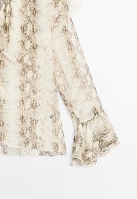 Blus med långa ärmar i genomskinligt tyg som har ett ormskinnsmönster i beige och bruna nyanser, med volanger vid ärmsluten och halsringningen.