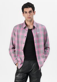 Camisa a cuadros gris y rosa con botones, de mangas largas, fabricada en un tejido suave y con un sutil logo en el lado izquierdo del pecho. Llevada sobre una blusa negra.