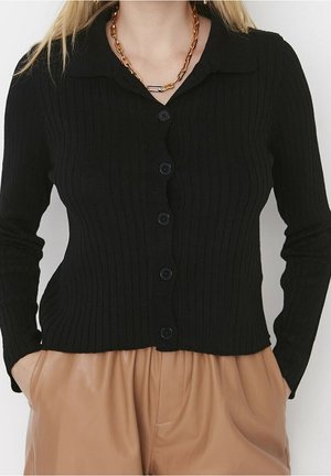 Cardigan noir en tricot à côtes avec un col, fermeture à boutons sur le devant et manches longues, associé à un pantalon plissé tan.