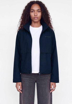 Un modèle se tient devant un fond blanc, vêtu d’une veste à capuche bleu marine foncé, d’une chemise blanche et d’un jean anthracite.