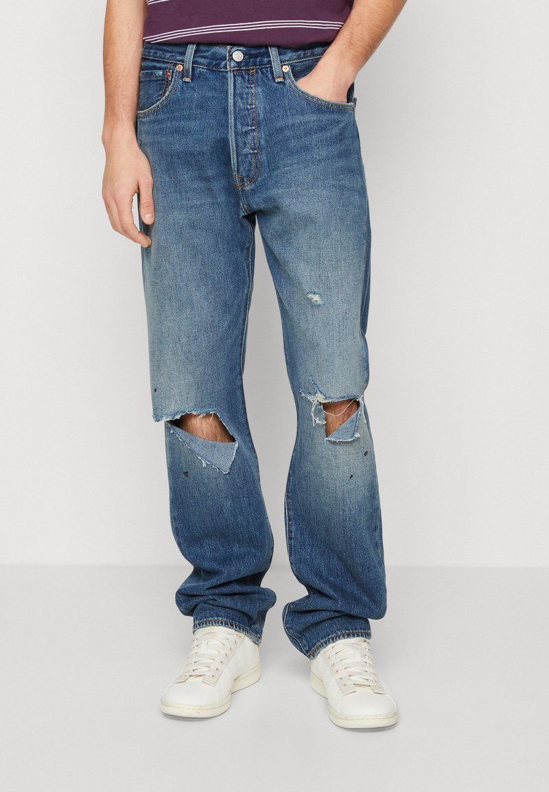 Jeans bleu clair avec une coupe décontractée, présentant de grandes déchirures aux genoux et un léger dégradé. Portés avec des baskets blanches.