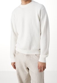 Felpa bianca a texture con motivo a rombi, con collo e polsini a costine, indossata con pantaloni di colore chiaro.