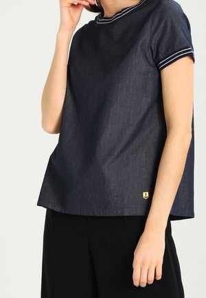 Nainen pukeutunut tummansiniseen, valkoisilla raidoilla koristeltuun lyhythihaiseen puseroon ja mustiin housuihin, käsi lähellä kasvojaan.