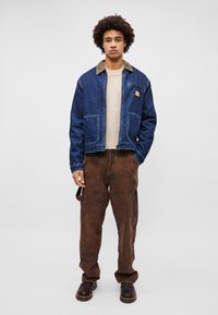 Jachetă din denim cu guler din catifea maro, două buzunare frontale și un patch. Purtați peste un pulover deschis și asortați cu pantaloni cargo maro și cizme.