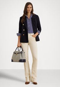 Blazer azul-marinho com botões dourados, camisa de botões às riscas, calças creme com corte evasê e uma mala cinzenta com detalhes azul-marinho. Sapatos castanhos de salto completam o visual.