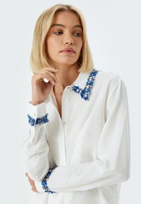 Camisa blanca con bordados florales azules en el cuello y los puños. Presenta un cierre frontal con botones y una tela texturizada.