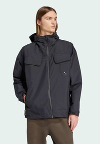Dark gray waterproof jacket with a zip closure, two large chest pockets, and a drawstring hood, made from lightweight fabric.