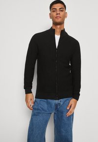 Svart zip-up cardigan med ribbet tekstur og ståkrage, båret over en hvit t-skjorte, parret med lyseblå jeans.