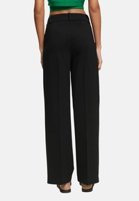 Femme portant un pantalon noir taille haute à jambes larges et un top court vert, debout dos à la caméra sur fond blanc.