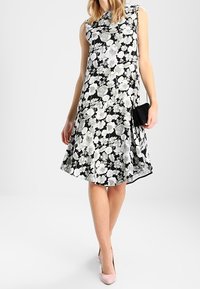 Žena v bezramenných černých šatech s bílými a šedými květinovými vzory, nese malou černou tašku přes rameno a růžové podpatky.