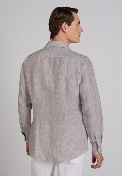 Hombre con camisa de manga larga beige y blanca a rayas finas y pantalones blancos, de espaldas contra un fondo liso gris claro.