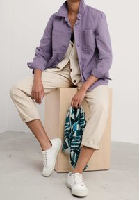 Lawendowa bawełniana kurtka z dwoma przednimi kieszeniami, zestawiona z kremowym prążkowanym topem i beżowymi, skróconymi spodniami. Białe trampki i wzorzysty szalik.