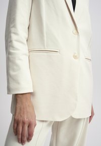 Krémový tailored blazer vyrobený z měkkého materiálu, s dvěma knoflíky, bočními kapsami a mírně zakřiveným lemem.