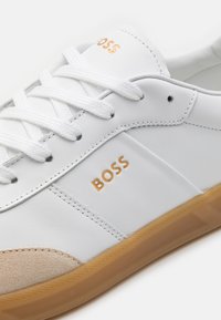 White leather sneaker with beige suede accents, golden "BOSS" branding, and white laces. Rubber sole with textured detail.