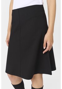Personne portant une jupe noire évasée jusqu'aux genoux, un haut blanc et des chaussettes montantes noires, avec une main posée à côté de la jupe.
