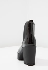 Černá kotníková bota z lakované kůže s mohutným podpatkem, opatřená elastickými bočními panely a texturovanou gumovou podrážkou. Hladký povrch s detaily švů.