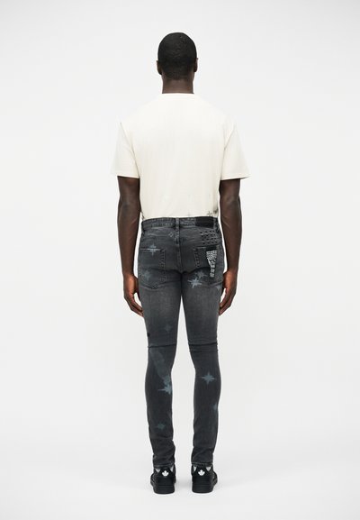 Άνδρας που φοράει λευκό κοντομάνικο πουκάμισο και σκουρόχρωμο γκρι στενό τζιν με αστέρια και διακοσμητικές λεπτομέρειες με τρουκς στην πίσω τσέπη, στέκεται με την πλάτη προς τα εμπρός.