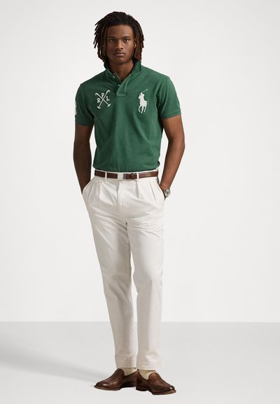 Grønn polo skjorte med hvit brodert logo, sammen med kremfargede bukser og brune slip-on sko. Modellen står med hendene i lommer.