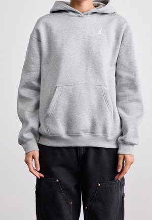 Person trägt einen hellgrauen Kapuzenpullover mit einer Fronttasche und schwarze Cargo-Hosen vor einem schlichten Hintergrund.