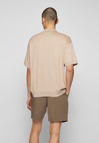 Beige polo tričko s krátkým rukávem a texturovanou tkaninou, s žebrovaným lemem, kombinované s hnědými šortkami, viděno zezadu.