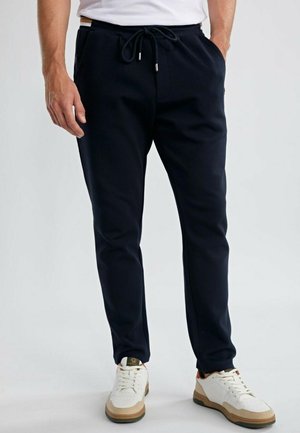 Homme portant un pantalon noir à cordon et des baskets blanches, debout avec une main dans la poche devant un fond uni.