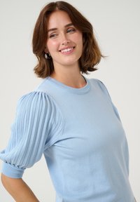 Un pull en tricot bleu clair avec un col rond et des manches bouffantes plissées. Le tissu semble doux avec une texture lisse.