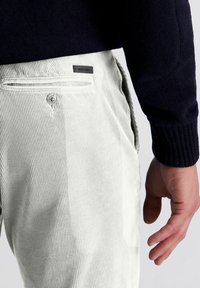 Witte corduroy broek met verticale ribbels, voorzien van een achterzak en een klein zwart logo label. Gedetailleerde stiksels en een gladde textuur zichtbaar.