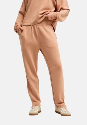 Personne portant un pantalon de survêtement beige coupe décontractée et un sweat assorti, les mains dans les poches, assorti de baskets blanches.