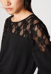 Haut noir avec un corps uni et des manches en dentelle transparente à motifs floraux. Encolure ronde et coupe décontractée, mettant en valeur un tissu texturé.
