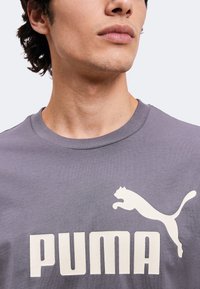 Νέος άνδρας που φοράει γκρι μπλούζα Puma με λευκό λογότυπο Puma και κείμενο, περικομμένο κάτω από τη μύτη και πάνω από το στήθος.