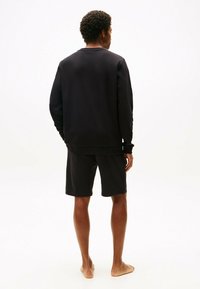 Completo con felpa nera e pantaloni corti. Tessuto liscio, vestibilità ampia. Design semplice senza loghi o motivi visibili. Posizione a piedi nudi.