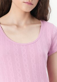 Jeune femme portant un haut rose clair texturé à manches courtes avec un décolleté arrondi, laissant voir le cou et la partie supérieure de la poitrine.