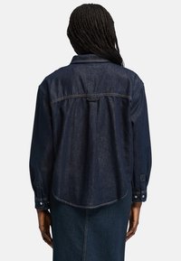 Camisa de mezclilla azul oscura con un ajuste relajado, cuello puntiagudo, mangas largas y dobladillo curvo. Presenta puños con botones y detalle de costura en la sisa trasera.