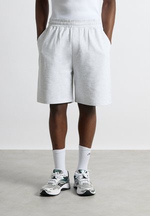 Person iført lysegrå knælange shorts, hvide ribstrikkede sokker og hvide og grønne sportssko, der står på en grå gulv.