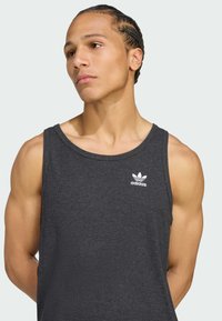 Tricou gri, cu dungi, fabricat din bumbac. Prezintă un gât rotund și logo-ul alb Adidas în partea stângă sus. Design fără mâneci.