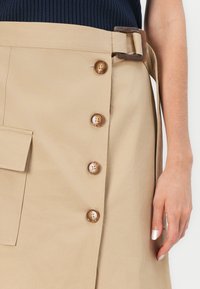 Beige skjørt i myk stoff med sidelomme, fem brune knapper og et belte med rektangulært spenne, med rett snitt og minimalistisk design.