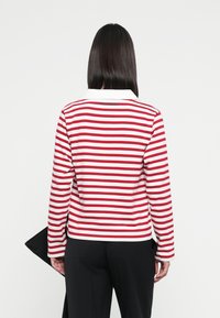 Langarmshirt mit rot-weißen horizontalen Streifen und einem weißen Kragen. Trägt schwarze Hose. Teilweise sichtbare schlichte schwarze Handtasche.