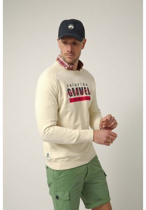 Hombre con gorra negra, sudadera beige con texto "veloride GRAVEL", camisa de cuadros roja debajo y pantalones cortos cargo verdes, ajustándose la manga.