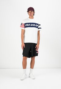 White t-shirt with navy "USA SAILGP" text, red and white striped sleeves; black shorts; white sneakers; maroon cap. Smooth fabric, casual fit.