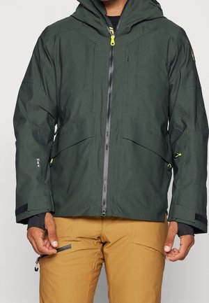 Mężczyzna ubrany w ciemnozieloną bluzę z kapturem i zamkiem z przodu oraz beżowe spodnie, ręce swobodnie luźno wzdłuż ciała na jasnym tle.