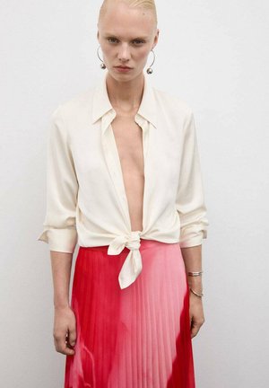 Crème zijde blouse geknoopt in de taille, gecombineerd met een geplooide rok die overgaat van roze naar rood. Grote zilveren creolen zichtbaar.