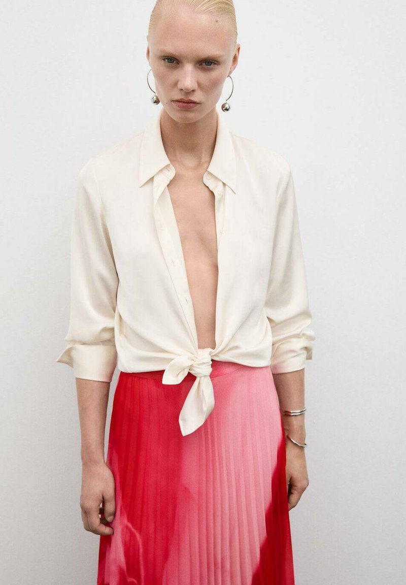 Crème zijde blouse geknoopt in de taille, gecombineerd met een geplooide rok die overgaat van roze naar rood. Grote zilveren creolen zichtbaar.
