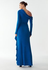 Naiste  ühesõlgne pikk sinine kleit, millel on langev varrukas ja hõbedased kõrged kontsad, seisab ühtlase valge taustal.