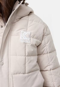 Beige vadderad jacka med quiltat mönster, med en logotypmärke på bröstet, knäppning med knappar och en huva för extra värme.