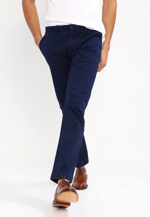 Mann geht, trägt dunkelblaue Hose, braune Lederschuhe und ein weißes T-Shirt mit den Händen in den Taschen.