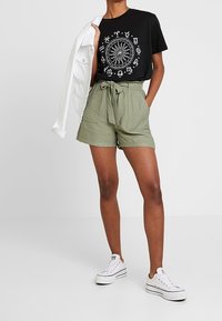 Černé tričko s bílým motivem slunce, olivově zelené šortky s poutky na pásek a zavazovacím pásem, a bílé tenisky. Jednoduchý, ležérní outfit.