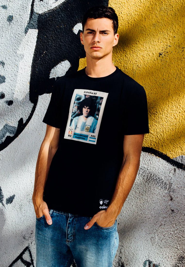 Zwarte katoenen T-shirt met een gedrukt ontwerp van een voetbalspeler kaart met het label "ESPAÑA 82." Ronde hals en korte mouwen, gecombineerd met vervaagde denim jeans.