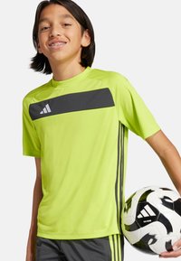 Camiseta atlética verde neón con una franja horizontal negra y un logo blanco, acompañada de pantalones cortos grises, sosteniendo un balón de fútbol en blanco y negro.