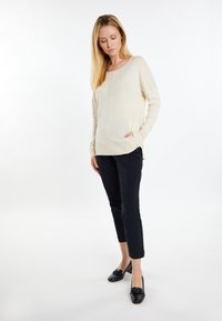 Beige Strickpullover mit seitlichen Schlitzen und langen Ärmeln, kombiniert mit maßgeschneiderten schwarzen Hosen und schwarzen Slippern. Glatte Textur über das gesamte Kleidungsstück.