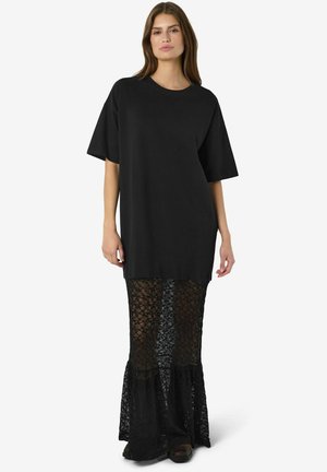 Žena nosi čierny oversized krátky rukáv top v kombinácii s dlhou, polopriehľadnou čiernou čipkovanou sukňou a čiernymi topánkami na bielom pozadí.